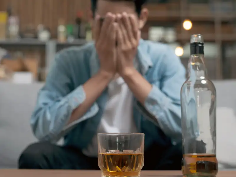 A man, covering his face, sat with a bottle of whisky in front of him