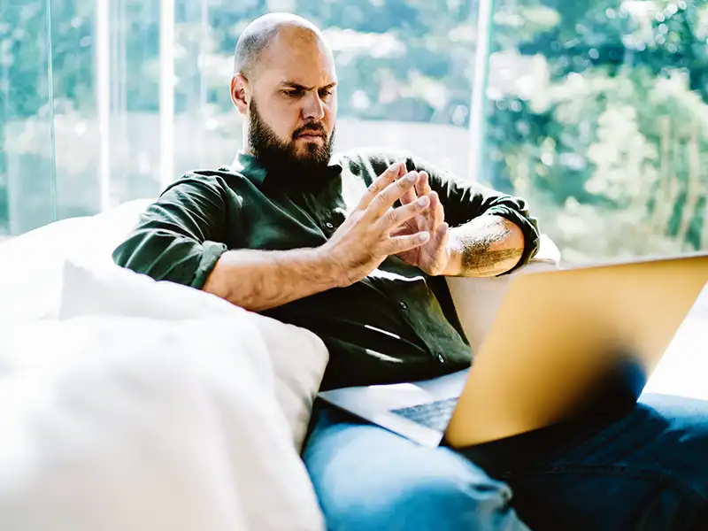 Stern man, in his living room, looking at his laptop with his hands together