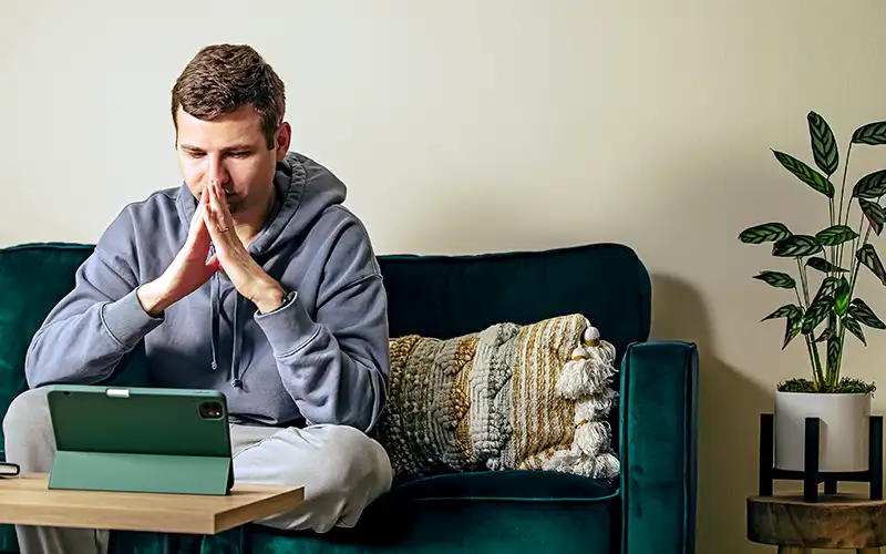 Young man, in his living room, looking at his tablet with his hands together